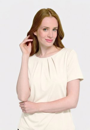 Junge Frau mit heller Haut und langen roten Haaren, die ein cremefarbenes Kurzarmoberteil trägt, berührt sanft ihr Haar und lächelt vor weißem Hintergrund.