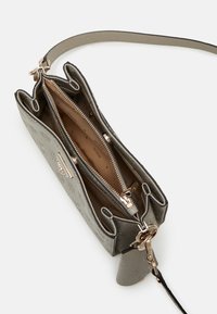Sac bandoulière gris avec motif de logo embossé, fermeture éclair dorée et fermoir, comprenant trois compartiments et une bandoulière amovible avec une petite pochette.