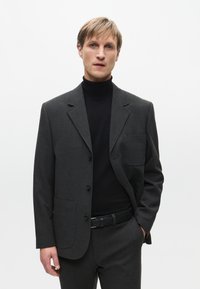 Blazer grigio con un design aderente, tasca sul petto singola e due tasche frontali. Indossato sopra un dolcevita nero, caratterizzato da una texture liscia.