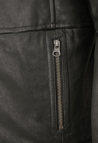 Schwarze Lederjacke mit strukturiertem Finish, sichtbaren Nähten und einem silbernen Reißverschluss an der vorderen Tasche.