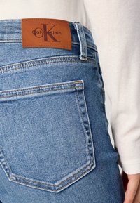 Plave traperice visokog struka, s smešnim kožnim logom Calvin Klein i ušivenim stražnjim džepovima. Vidljivi detalji teksturirane tkanine.