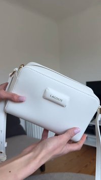 Personne tenant un sac bandoulière blanc Lacoste avec une fermeture éclair et une sangle dorées, à l’intérieur avec un fond neutre.