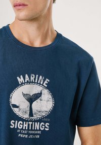 Camiseta de algodón azul marino con un gráfico de cola de ballena y un diseño de texto circular en blanco. Cuello redondo acanalado con mangas cortas.
