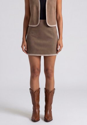 Donna con mini gonna in camoscio marrone, gilet abbinato con rifinitura bianca, top nero e stivali da cowboy in pelle marrone che sta davanti a uno sfondo semplice.