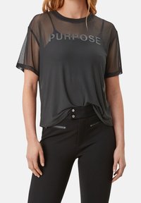 Sort mesh t-shirt med "PURPOSE" i grå skrift. Har korte ærmer og en løstsiddende pasform, parret med sorte bukser med lynlåselementer.
