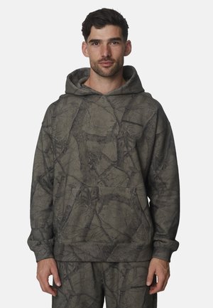 Uomo che indossa una felpa con cappuccio grigio-verde e pantaloni abbinati con un delicato motivo mimetico di foglie e rami, in piedi davanti a uno sfondo semplice.