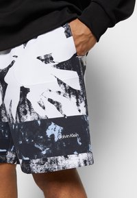 Shorts noir et blanc ornés de motifs de feuilles abstraites et d'un accent bleu. Le tissu semble léger avec un logo sur le bas de l'ourlet.