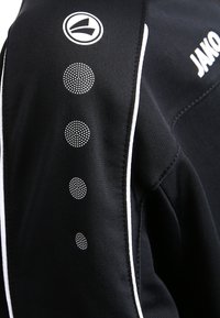 Zwarte blazer met sportieve uitstraling, voorzien van witte accenten, kleine gestipte patronen op de schouder en een logo op de kraag. Gemaakt van een textuurstof.