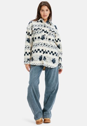 Chemise en polaire crème avec des motifs géométriques et floraux bleu marine. Dotée d'une fermeture à boutons, de deux poches poitrine et d'une coupe décontractée. Associée à un jean à jambes larges.