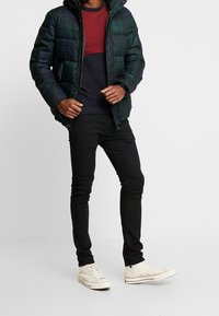 Vadderad grön och svart rutig jacka med huva, bärs över en röd och marinrandig tröja, kombinerad med svarta skinny jeans och beige sneakers.