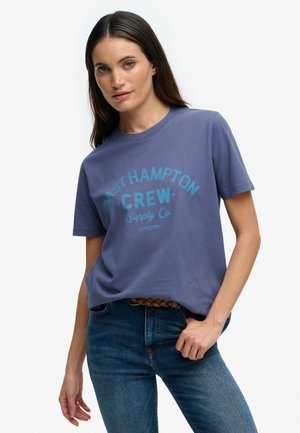 Femme aux longs cheveux foncés portant un t-shirt bleu « East Hampton Crew Supply Co. » et un jean bleu avec une ceinture tressée, debout devant un fond blanc.