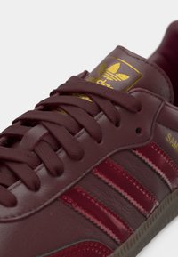 Αθλητικό παπούτσι από δέρμα μπορντό με χρυσές λεπτομέρειες, υφασμένα κορδόνια και τρεις γυαλιστερές ρίγες. Διαθέτει λεία υφή και λαστιχένια σόλα.