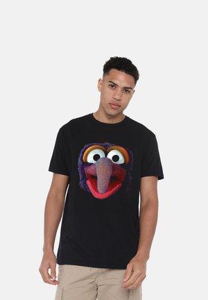 Junger Mann trägt ein schwarzes T-Shirt mit einem bunten Cartoon-Vogelgesicht auf der Vorderseite und steht vor einem schlichten hellen Hintergrund.
