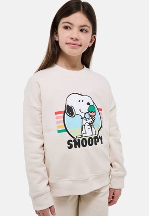 Fille portant un sweat-shirt beige clair avec Snoopy tenant un cornet de glace coloré et des rayures arc-en-ciel en arrière-plan.
