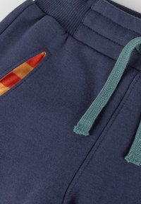 Marineblaue Sweatpants mit einem elastischen Bund, kontrastierendem petrolfarbenem Kordelzug und orange-gelbem karierten Innenfutter der Taschen.
