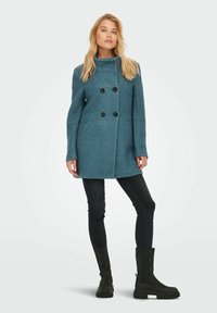 Teal dubbelzijdig jasje met hoge kraag, zwarte knopen en voorzakken; model draagt skinny zwarte jeans en zwarte kniehoge laarzen.