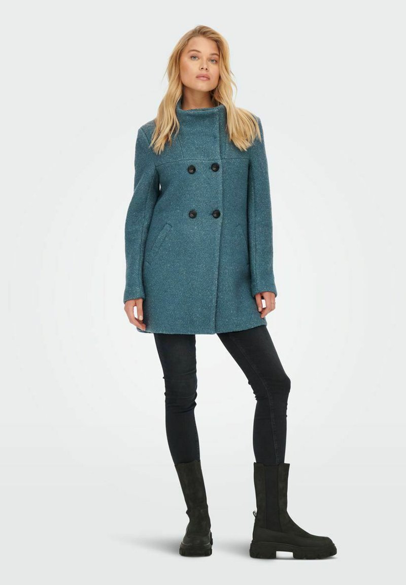 Tee-farbener doppelter Mantel mit hohem Kragen, schwarzen Knöpfen und vorderen Taschen; das Modell trägt enge schwarze Jeans und schwarze kniehohe Stiefel.