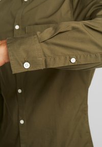 Man klädd i en olivgrön långärmad skjorta med vita knappar, ärmupphängd med knapp, armen sträckt utåt.