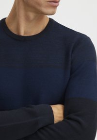 Temno modra pletena majica z okroglim izrezom. Ima teksturiran vzorec s horizontalnimi črtami. Rebraste manšete in rob.