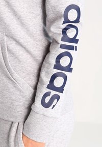 Sweat-shirt gris à texture douce avec un grand logo Adidas bleu marine imprimé verticalement sur la manche gauche. Comprend une poche avant.
