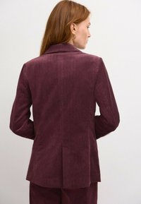Burgundin värinen corduroy-takki, jossa on herringbone-kuvio, yksittäinen ventti, ja lovikaulukset, joissa on pitkät hihat ja takataskuyksityiskohta.