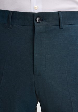 Gros plan sur un pantalon formel bleu foncé avec une taille boutonnée et des passants de ceinture, porté avec une chemise blanche.
