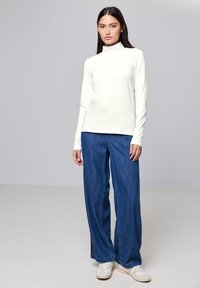 Witte turtleneck met lange mouwen, gedragen bij ruime, donkerblauwe spijkerbroeken en witte sneakers. Eenvoudig ontwerp met een gladde textuur.