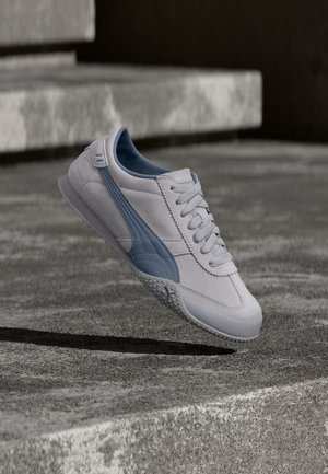 Lysegrå og blå Puma-sneaker med snørebånd, hængende over teksturerede betontrin i udendørs omgivelser.