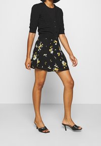 Zwarte gebreide cardigan met korte mouwen, met knoopdetails, gecombineerd met een zwarte rok met bloemenpatroon met gele en witte bloemen. Zwarte hakken sandalen.