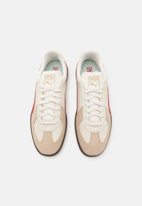 Svetle béžové a bílé tenisky s červenými akcenty, hladké a texturované povrchy, šněrovací design a hnědá gumová podrážka. Na jazyku je umístěno logo.