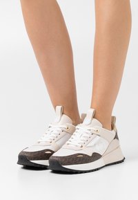 Lichtbeige sneakers met bruine accenten, voorzien van een textuurpatroon, gouden oogjes en een iets verhoogde zool.