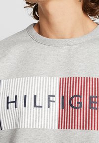 Felpa grigia con strisce orizzontali bianche, blu marino e rosse, con il testo "HILFIGER" in blu marino al centro. Maniche a girocollo.
