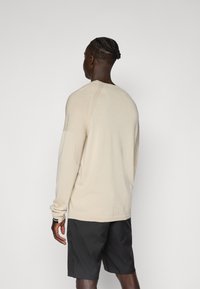 Beige Pullover mit langen Ärmeln und strukturierten Ärmeln, lockerer Passform und gerippten Details am Kragen und Saum, kombiniert mit dunklen Shorts.