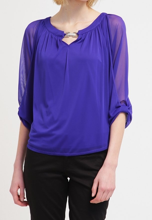 Vrouw draagt een koningsblauwe blouse met doorschijnende driekwart mouwen en een zilveren gespdetail bij de halslijn, gecombineerd met zwarte broek.