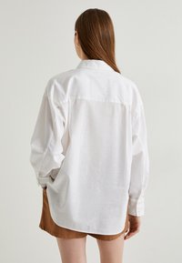 Hvid linnedskjorte med lange ærmer, knapmanchetter og en afslappet pasform. Har en rygfældning og rund kant. Kombineret med brune shorts.
