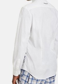 Zadní pohled na osobu, která má na sobě bílé dlouhé rukávy tričko s nápisem u límce a modré kostkované kalhoty.