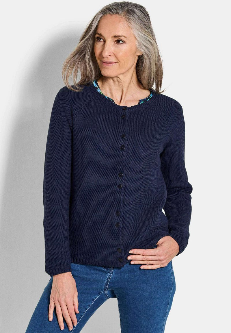 Frau mit grauem Haar, die eine navyblaue Knopfleiste und blaue Jeans trägt, steht mit einer Hand auf der Hüfte vor einem schlichten Hintergrund.