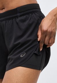 Κοντινό πλάνο ατόμου που φοράει μαύρο διπλό στρώμα αθλητικών σορτς, χέρι που ρυθμίζει τη σχισμή στο εξωτερικό στρώμα κοντά στον μηρό, με το λογότυπο της Asics εμφανές στα σορτς.