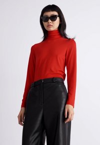 Haut turtleneck rouge à manches longues associé à un pantalon en cuir noir taille haute. Le mannequin porte des lunettes de soleil noires. Tissu lisse et ajusté.