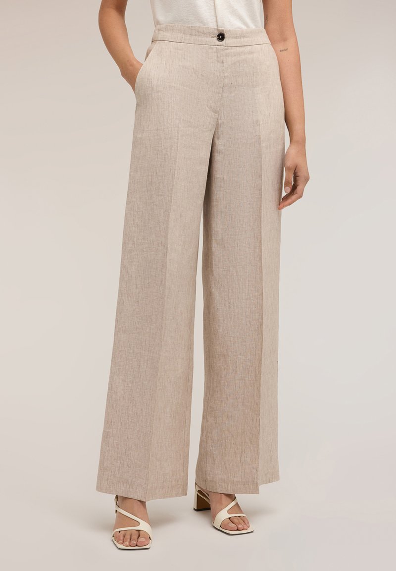 Mujer con pantalones de lino beige de pierna ancha con pliegue frontal, bolsillos laterales, cinturilla con botones y sandalias blancas de tacón.