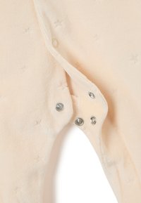 Mameluco de color durazno con tela suave y texturizada. Presenta cierres a presión en la parte inferior y un sutil patrón de estrellas en toda su superficie.