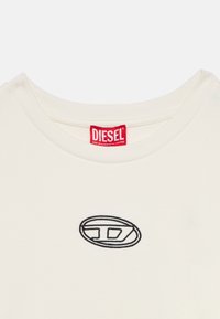 Μπεζ βαμβακερό μπλουζάκι με στρογγυλή λαιμόκοψη. Διαθέτει μαύρο κεντητό λογότυπο και κόκκινη ετικέτα με λευκό κείμενο.