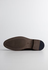 Sohle eines braunen Leder-Dressschuhs, flaches Design, strukturierter Gummi für Grip, mit subtiler Markenkennzeichnung und einem kontrastierenden marineblauen Akzent an der Spitze.