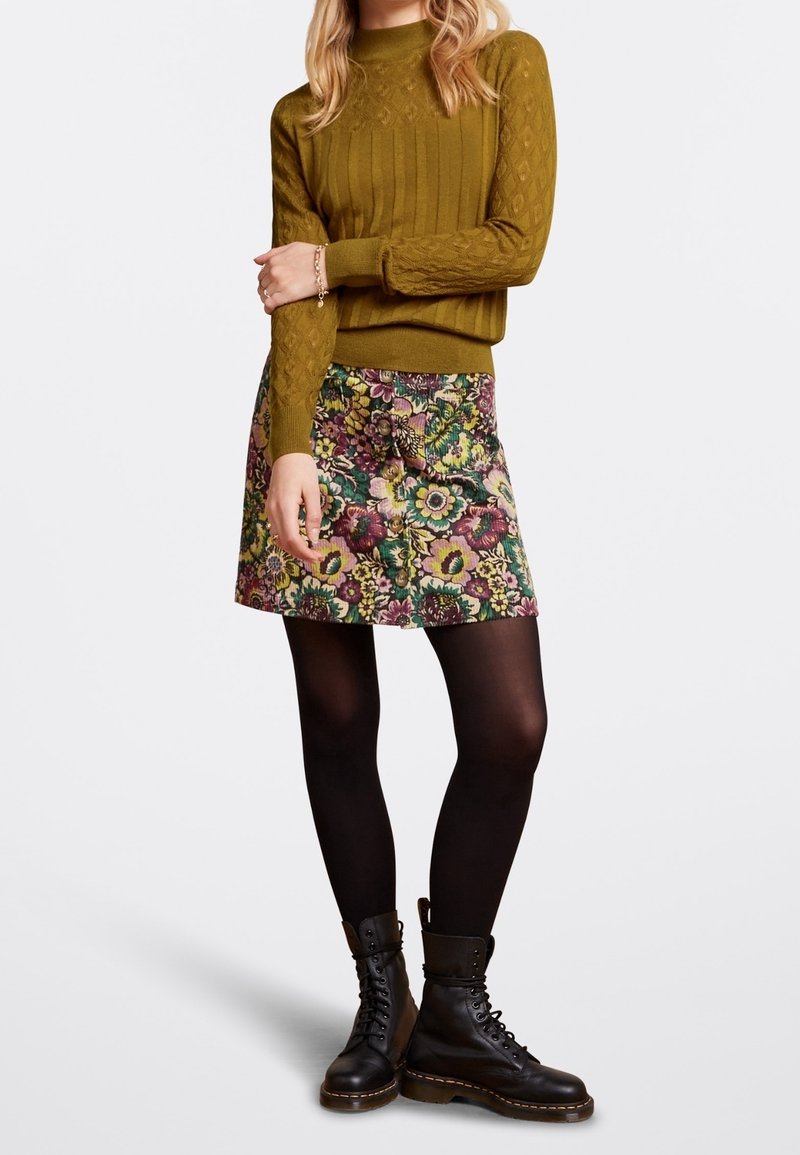 Femme portant un pull en maille torsadée vert olive, une jupe à motif floral, des collants noirs et des bottes noires à lacets, debout les bras croisés.
