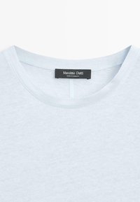 T-shirt à col ras du cou bleu clair avec une étiquette noire indiquant "Massimo Dutti, Fabriqué au Cambodge" cousue à l'intérieur du col.