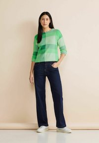 Zielony sweter w kratkę z długimi rękawami, zestawiony z ciemnoniebieskimi jeansami o szerokich nogawkach i białymi trampkami. Gładkie tło podkreśla widoczność.