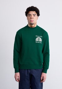 Sudadera verde con cuello redondo y mangas largas. Presenta un logo y texto bordados en blanco en el pecho. Textura suave, corte casual.