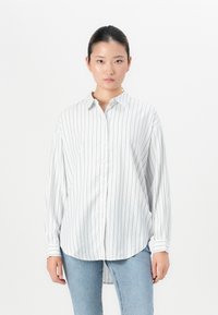Kvinne iført en hvit langermet skjorte med tynne svarte vertikale striper og lyseblå jeans, stående mot en ensfarget hvit bakgrunn.