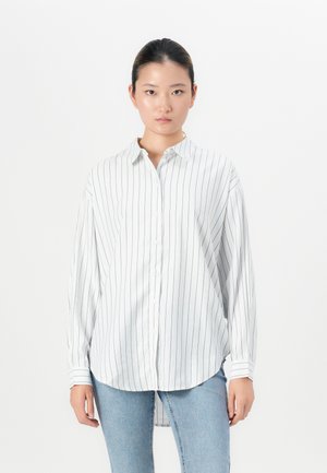 Woman wearing a white long-sleeve button-up shirt with thin black vertical stripes and light blue jeans, standing against a plain white background.