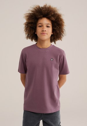 Ung person med krøllet hår iført en ensfarvet lilla t-shirt og mørke bukser, stående mod en neutral baggrund med hænderne bag ryggen.